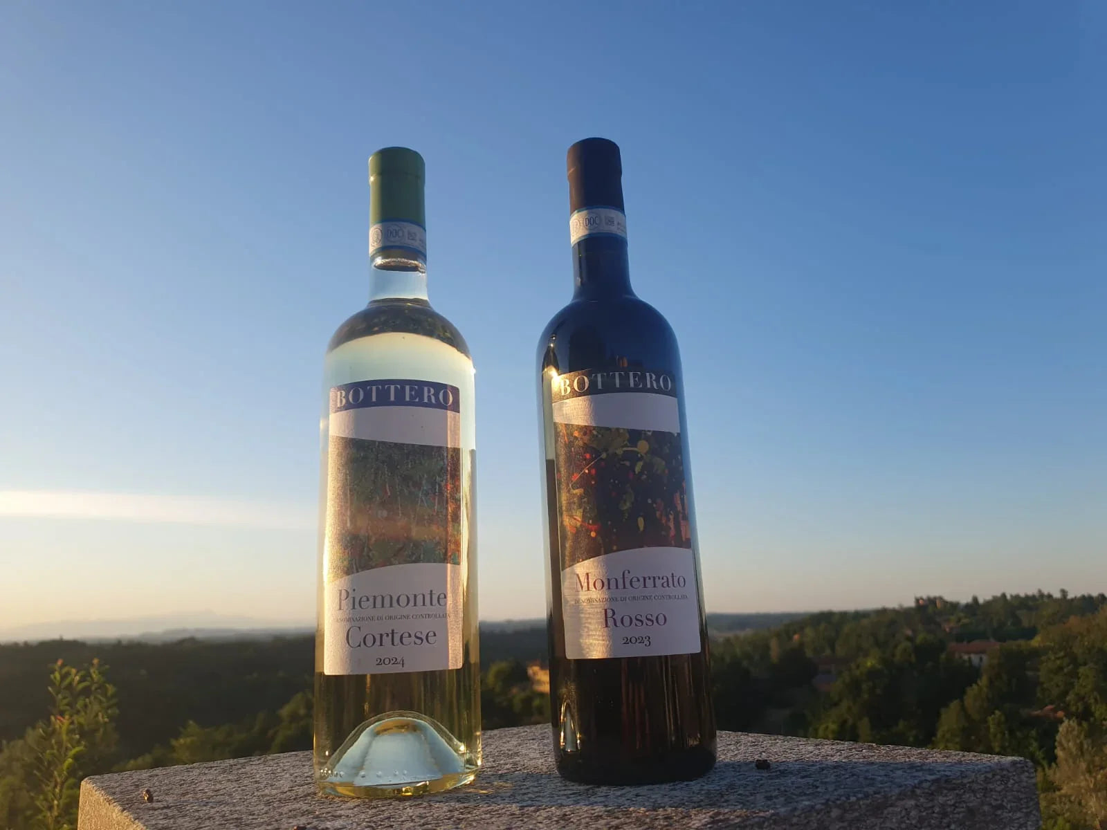 Two Bottero wine bottles, Cortese white and Monferrato Rosso red, displayed outdoors at sunset.